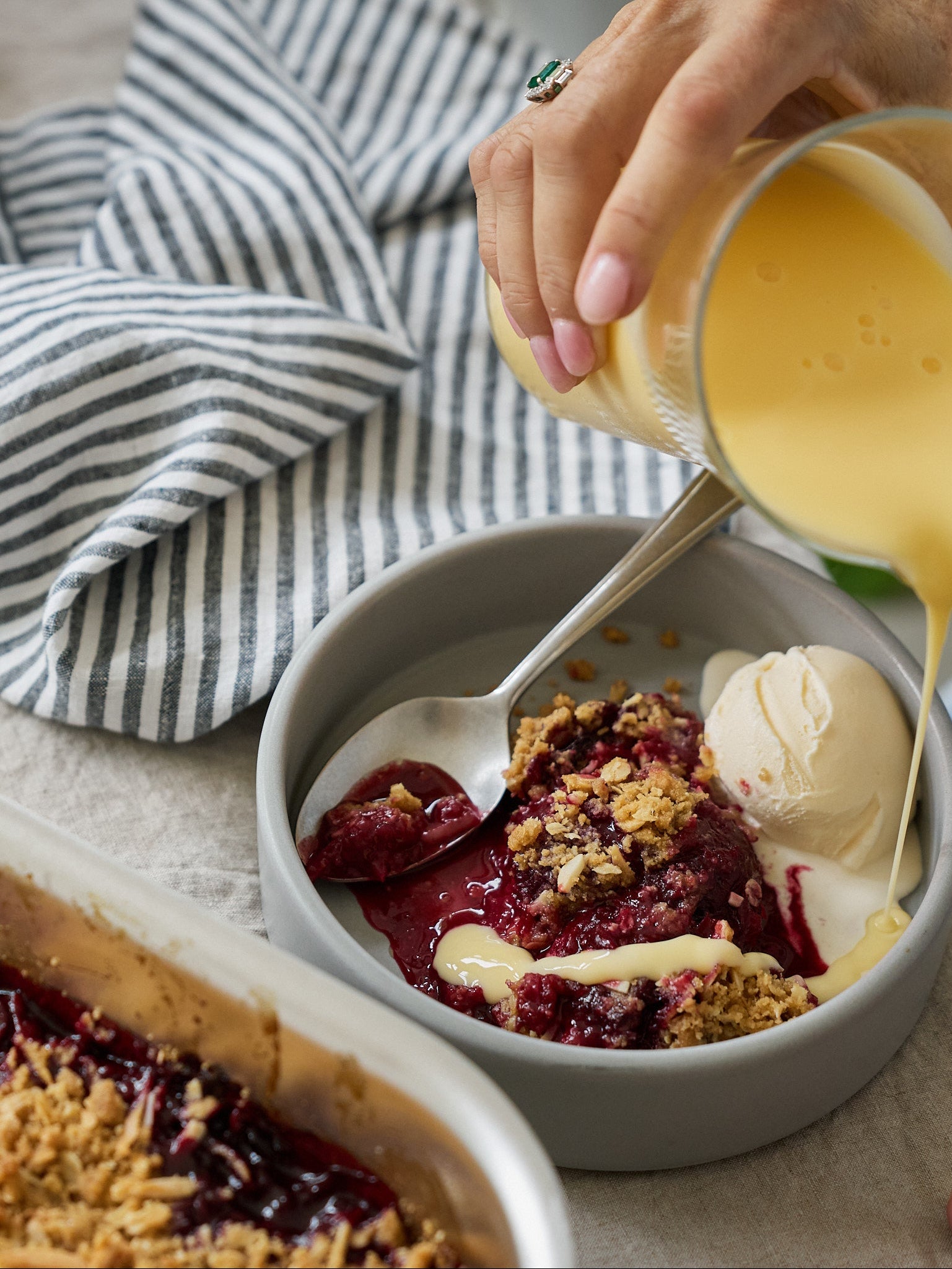 Person pouring custard over a dessert in a bowl with a striped napkin and another dessert in the background.
